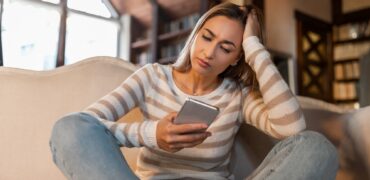 Bad News Concept. Upset Confused Woman Holding Smartphone, Looking At Mobile Phone Screen With Worried Expression, Touching Head, Sad Adult Female Reading Unplesant Message Sitting On Couch